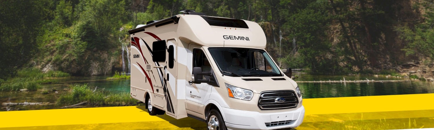 White & red-striped 2019 Gemini Class C motorhome above a lake surrounded by trees.